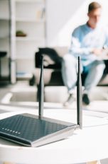Ein Router steht auf einem weißen, runden Tisch im Vordergrund. Dahinter sitzt eine Person auf einem Sessel und bedient ein Tablet.