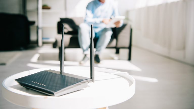 Ein Router steht auf einem weißen, runden Tisch im Vordergrund. Dahinter sitzt eine Person auf einem Sessel und bedient ein Tablet.