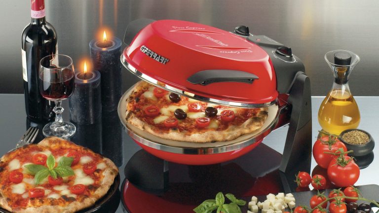 Auf einem Tisch neben einer selbstgebackenen Pizza, einem Rotwein, einem Paar Kerzen und diversen Pizzazutaten steht ein halb geöffneter Pizzaofen von Delizia, in dem eine weitere fertige Pizza liegt.