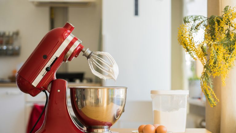 Eine KitchenAid Küchenmaschine steht auf einem Tisch, am Schneebesen klebt etwas Teig. Vor der Maschine stehen Mehl und ein paar Eier in einer Schale.