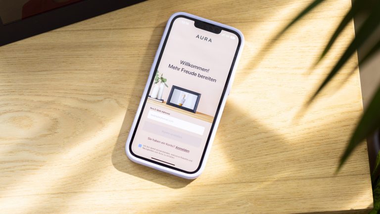 Ein Smartphone liegt auf einem Holzuntergrund. Darauf ist die App „AURA“ geöffnet. aura-bilderrahmen-9977