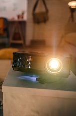 Beamer Ein Projektor steht auf einem kleinen Schränkchen in einem Wohnzimmer. Ein Laptop ist an ihn angeschlossen.