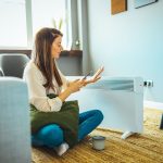 Eine Person sitzt vor einem Heizl&uuml;fter und schaut auf ihr Smartphone.