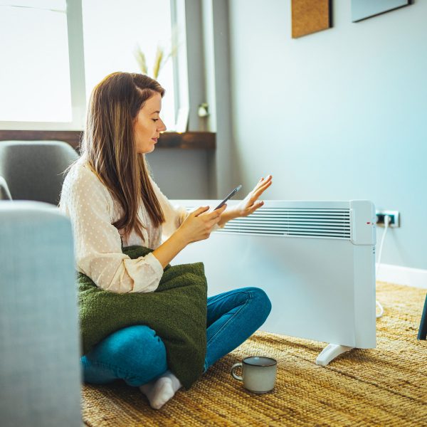 Eine Person sitzt vor einem Heizl&uuml;fter und schaut auf ihr Smartphone.
