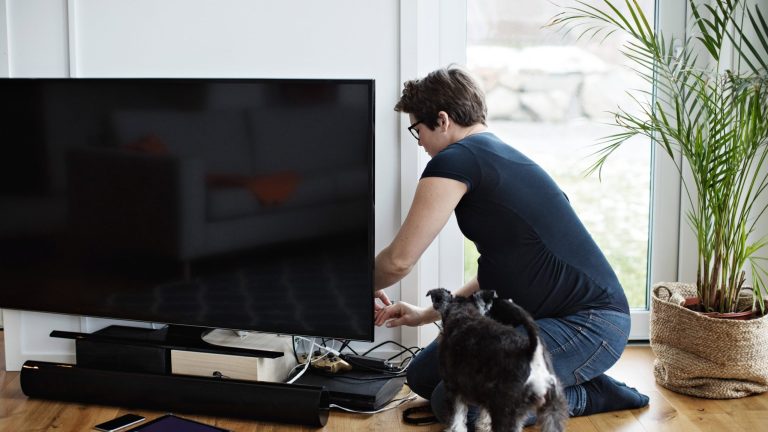 Eine Person kniet neben einem Fernseher und schließt einige Geräte per Kabel an diesen an.