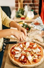 Eine Familie steht an einem Holztisch und belegt gemeinsam eine selbstgebackene Pizza.