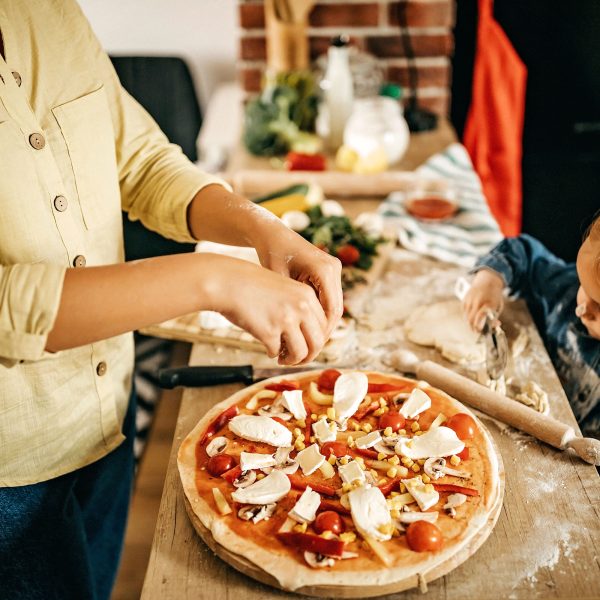 Eine Familie steht an einem Holztisch und belegt gemeinsam eine selbstgebackene Pizza.
