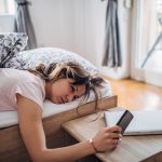 Eine m&uuml;de aussehende Person liegt im Bett und h&auml;lt ein Smartphone in der Hand.