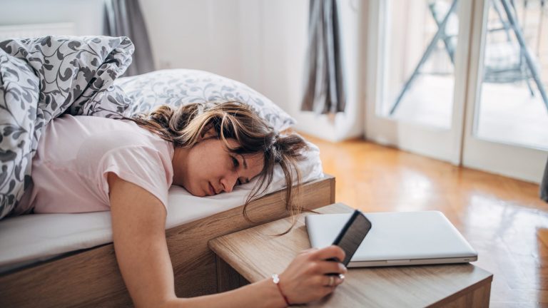 Eine müde aussehende Person liegt im Bett und hält ein Smartphone in der Hand.