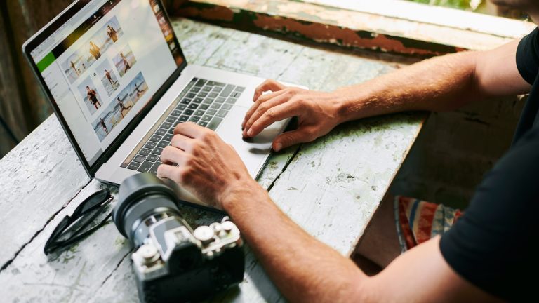 Eine Person sitzt vor einem Laptop und betrachtet Bilder. Daneben liegt eine Kamera.