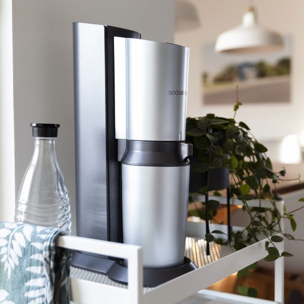 Ein silberner SodaStream steht auf einem wei&szlig;en Regal neben einer Pflanze und einer Glasflasche.