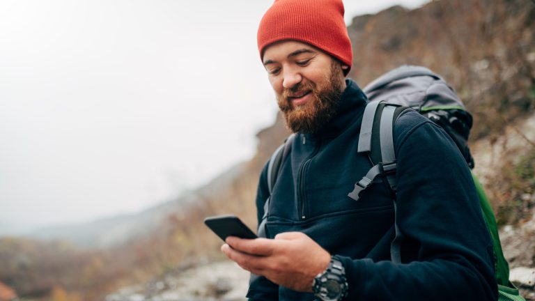 Eine in Wandergarderobe gekleidete Person in den Bergen schaut auf ein Smartphone.