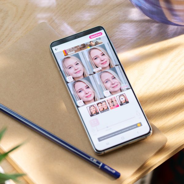 Ein Android liegt auf einem Holztisch. Auf dem Display ist ein Screenshot der App „YouCam Makeup“ zu sehen.