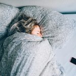 Eine Person liegt bis zur Nase in eine Decke gewickelt in einem Bett. Neben ihr liegt ein Smartphone.