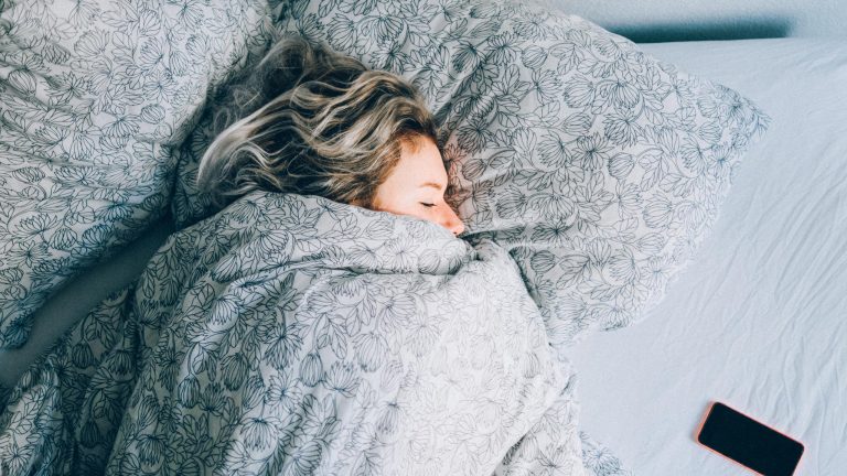 Eine Person liegt bis zur Nase in eine Decke gewickelt in einem Bett. Neben ihr liegt ein Smartphone.