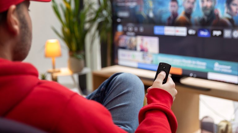 Eine Person sitzt vor einem Fernseher mit der Fernbedienung eines Fire-TV-Sticks in der Hand.
