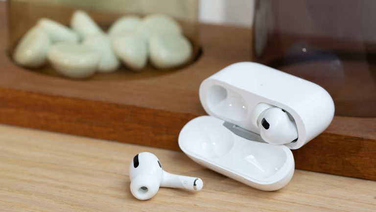 In einer weißen Ladebuchse befindet sich ein In-Ear-Kopfhörer. Der zweite liegt daneben auf einem Tisch.