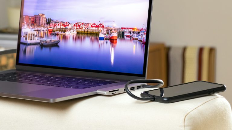 Neben einem MacBook liegt ein Smartphone, das mittels Kabel und Dockingstation mit dem Laptop verbunden ist.