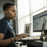 Eine Person sitzt an einem Schreibtisch, auf dem ein Laptop im Vordergrund und ein Monitor dahinter stehen.