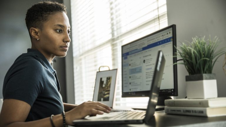 Eine Person sitzt an einem Schreibtisch, auf dem ein Laptop im Vordergrund und ein Monitor dahinter stehen.