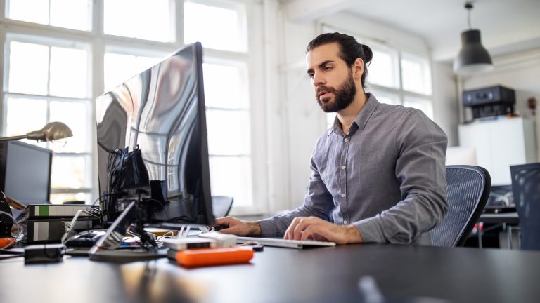 Eine Person sitzt vor einem Computer, Neben dem Bildschirm liegt eine externe Festplatte mit orangefarbiger Schutzhülle.