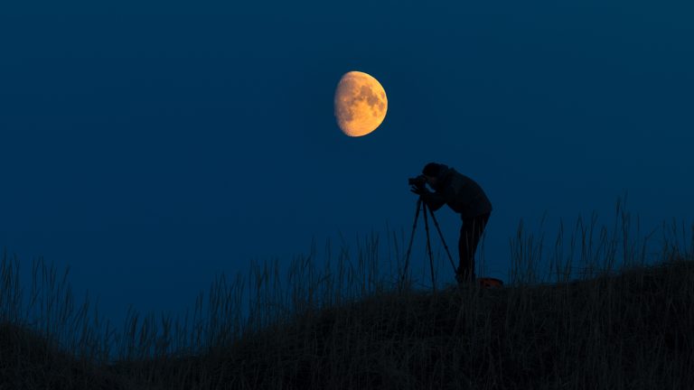 Der in Orange gehüllte Mond wird von einer Person mit Stativ fotografiert.