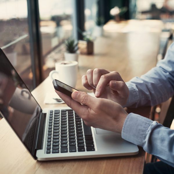 Eine Person sitzt in einem Caf&eacute; vor einem MacBook und bedient derweil ein Smartphone in der Hand.