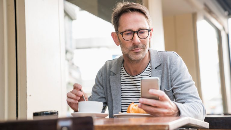 Eine Person sitzt bei Kaffee und Kuchen draußen an einem Tisch. In der einen Hand hält sie ein Smartphone.