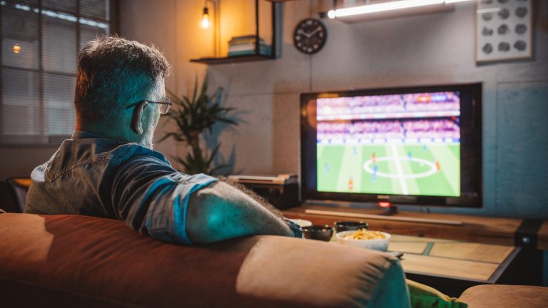 Eine Person sitzt auf einem Sofa und schaut ein Fu&szlig;ballspiel auf einem TV.