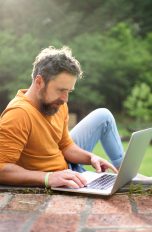 Eine Person sitzt, umringt von Blumen, draußen vor einem Laptop.