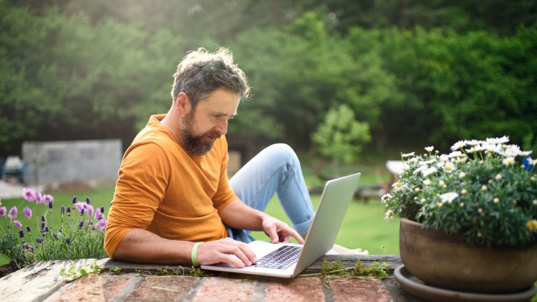 Eine Person sitzt, umringt von Blumen, draußen vor einem Laptop.