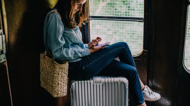 Eine Person sitzt in einem Zug auf ihrem Koffer und tippt auf dem Handy.