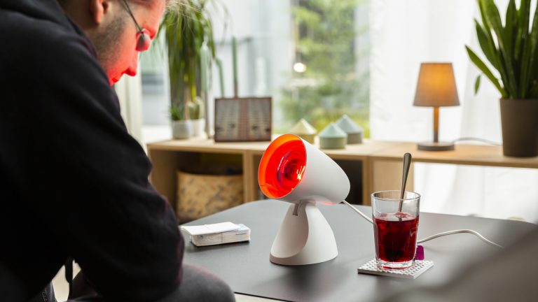 Eine Person sitzt vor einer Rotlichtlampe. Daneben steht eine Tasse Tee.