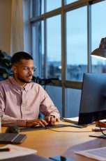 Eine Person sitzt an einem Schreibtisch vor einem Rechner. Neben dem Bildschirm steht eine Schreibtischlampe.