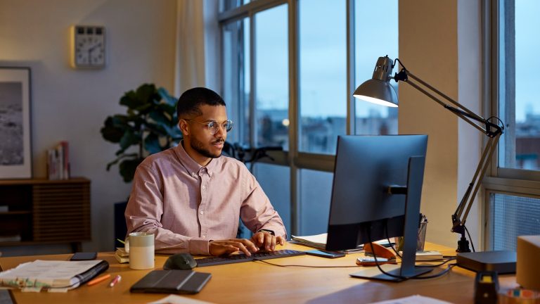 Eine Person sitzt an einem Schreibtisch vor einem Rechner. Neben dem Bildschirm steht eine Schreibtischlampe.