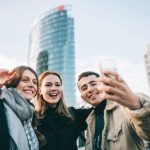 Drei Personen stehen vor hohen Geb&auml;udekomplexen und machen freudestrahlend ein Selfie.