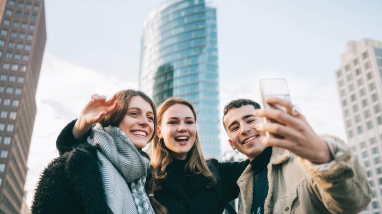 Drei Personen stehen vor hohen Gebäudekomplexen und machen freudestrahlend ein Selfie.