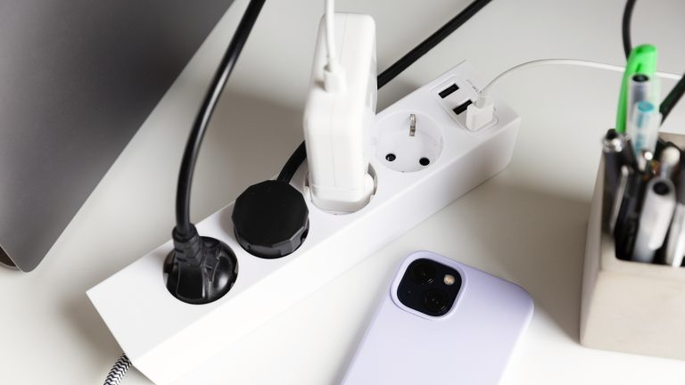 Eine Mehrfachsteckdose mit drei eingesteckten Ger&auml;ten und einem Smartphone-Ladeger&auml;t liegt auf einem Tisch.