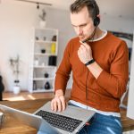 Eine Person sitzt auf einem Tisch und hat einen Laptop auf dem Scho&szlig;. Sie tr&auml;gt ein Headet auf dem Kopf.
