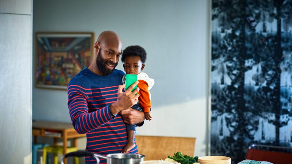 Eine Person mit einem Kind auf dem Arm steht vor einer Kücheninsel. In der freien Hand hält sie ein Smartphone, in das die beiden lächelnd hineinschauen.