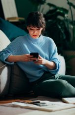 Eine Person sitzt vor einer Couch auf dem Boden und schaut auf ihr Smartphone.