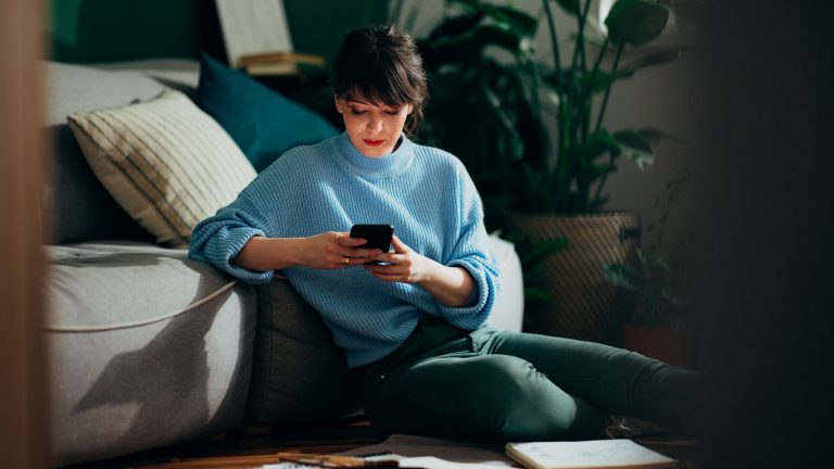 Eine Person sitzt vor einer Couch auf dem Boden und schaut auf ihr Smartphone.