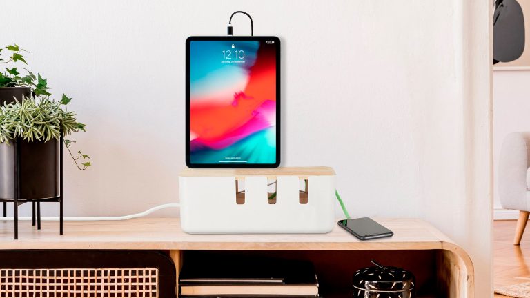 Ein iPad steht auf einer Kabelbox, die wiederum auf einem Schreibtisch aus hellem Holz gestellt wurde.