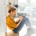 Eine Person lehnt sitzend an einer Wand in einem Wohnzimmer und schaut auf das Smartphone in ihren H&auml;nden.