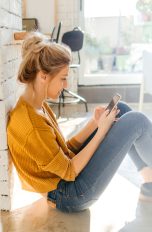 Eine Person lehnt sitzend an einer Wand in einem Wohnzimmer und schaut auf das Smartphone in ihren Händen.