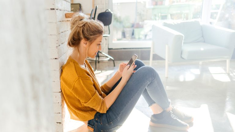 Eine Person lehnt sitzend an einer Wand in einem Wohnzimmer und schaut auf das Smartphone in ihren Händen.