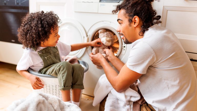 Ein Kind sitzt in einem Wäschekorb und gibt zusammen mit einer Person einen Stoffbären in die Waschmaschine.
