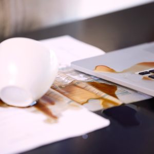 Eine weiße Kaffeetasse liegt samt Inhalt ausgebreitet auf Dokumenten und einer Laptop-Tastatur.