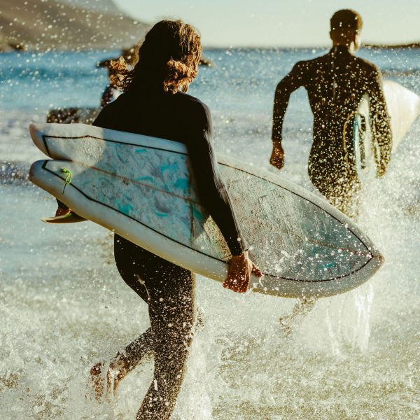 Personen mit Surfbrettern unter dem Arm und Neoprenanzügen laufen ins offene Meer.