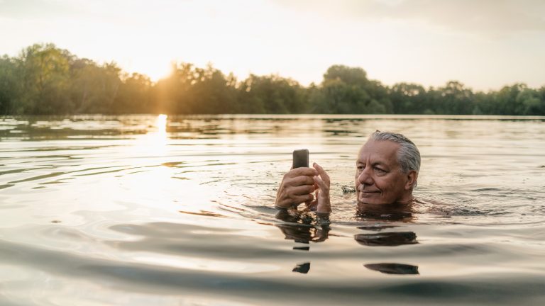 Eine Person schwimmt in einem See und bedient dabei ein Smartphone.